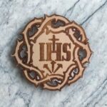Na nahrané fotografii je kruhový dřevěný magnet o průměru 10 cm, gravírovaný do světlé bukové překližky. Uprostřed motivu se nachází velký nápis IHS, který je křesťanskou zkratkou pro jméno Ježíš. Nápis obepíná trnová koruna, jejíž ostny směřují dovnitř a připomínají Kristovo utrpení. Pod nápisem jsou vyobrazeny tři hřeby, z nichž dva míří šikmo dolů z boků a jeden přímo dolů ze středu – odkazují na nástroje ukřižování.