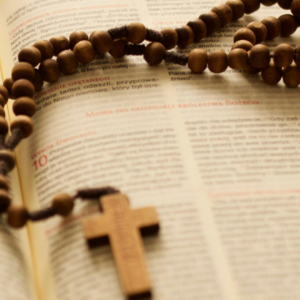 Detailní záběr na dřevěný růženec položený na otevřené Bibli. Kulaté dřevěné korálky jsou navlečené na tmavé šňůrce a vedou k jednoduchému dřevěnému kříži, který spočívá na stránce s textem Písma. Text v knize je mírně rozostřený, zatímco růženec je v popředí ostrý, což zdůrazňuje propojení modlitby růžence s Božím slovem. Celá scéna je laděna do teplých, jemných tónů a působí klidně a rozjímavě.