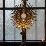 Zlatá paprsková monstrance před oknem vedle hořící svíčky