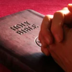 Sepjaté ruce položené vedle tmavé Bible na červené látce