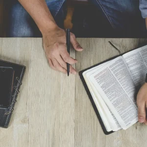 Otevřená Bible na stole a ruce zapisující poznámky