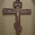 Historický dřevěný kříž s drobnými reliéfními výjevy na světlé stěně