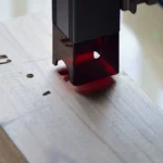Detail laserové hlavy pracující nad světlou dřevěnou deskou