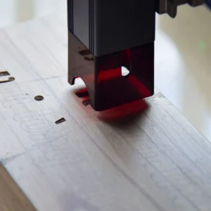 Detail laserové hlavy pracující nad světlou dřevěnou deskou