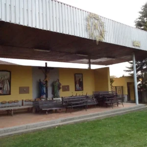 Venkovní poutní kaple s lavičkami a křížem pod širokou střechou